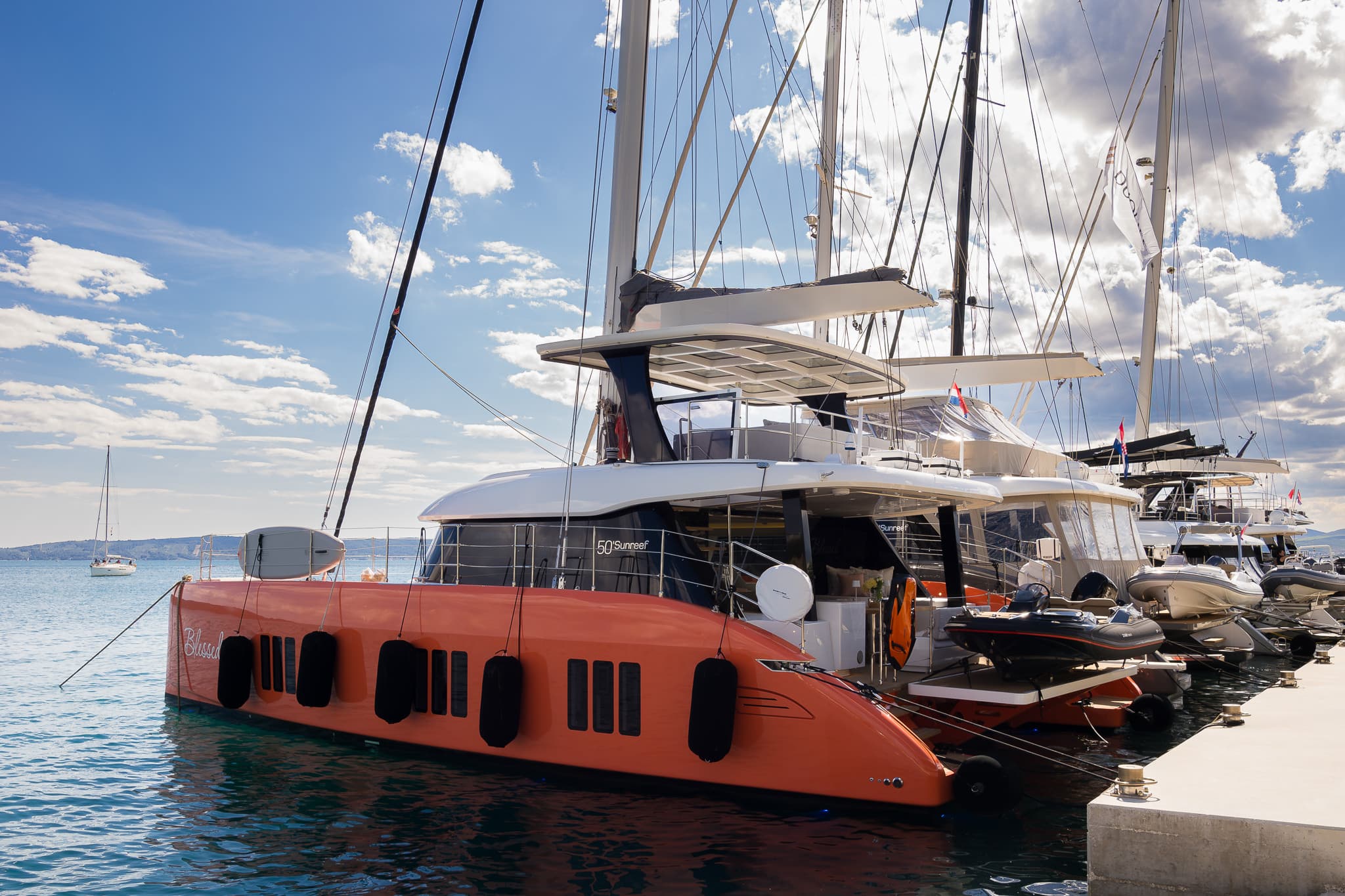 Luxuriöser Sunreef 50 Katamaran mit orangefarbenem Rumpf, festgemacht im Hafen unter blauem Himmel mit weißen Wolken