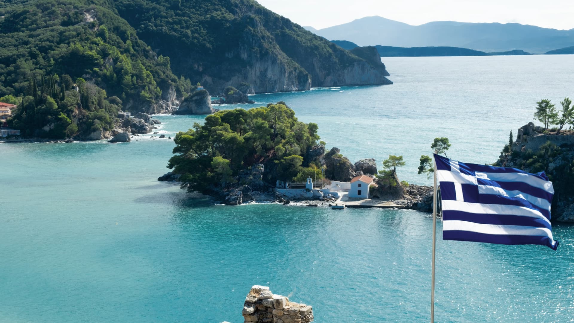 Griechische Insel mit Kapelle vor der Küste von Parga, türkisfarbenes Wasser, üppige grüne Vegetation und wehende griechische Flagge.