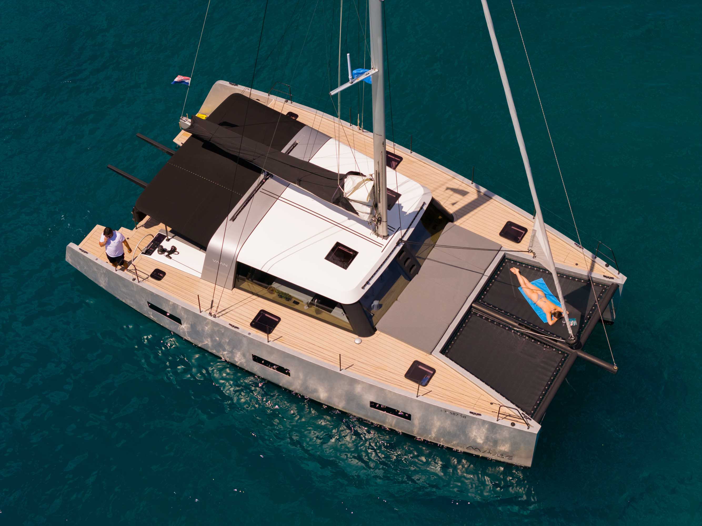 Luftaufnahme eines modernen Elektro-Katamarans VAAN R4 Silence vor Anker in türkisfarbenem Wasser vor Kroatien. Eine Person sonnt sich auf dem Trampolin, eine weitere bereitet das Deck vor.