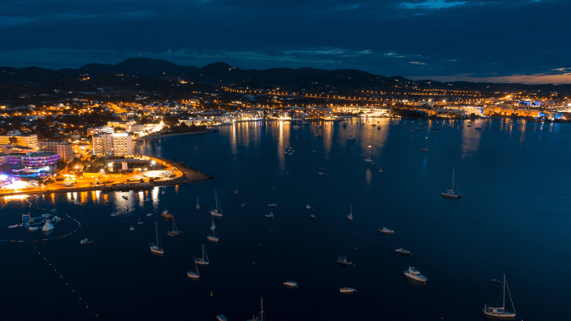 Glitzernde Lichter von Ibiza bei Nacht, gesehen vom Deck eines Katamarans – Ein unvergesslicher Moment während eines exklusiven FKK-Segeltörns.