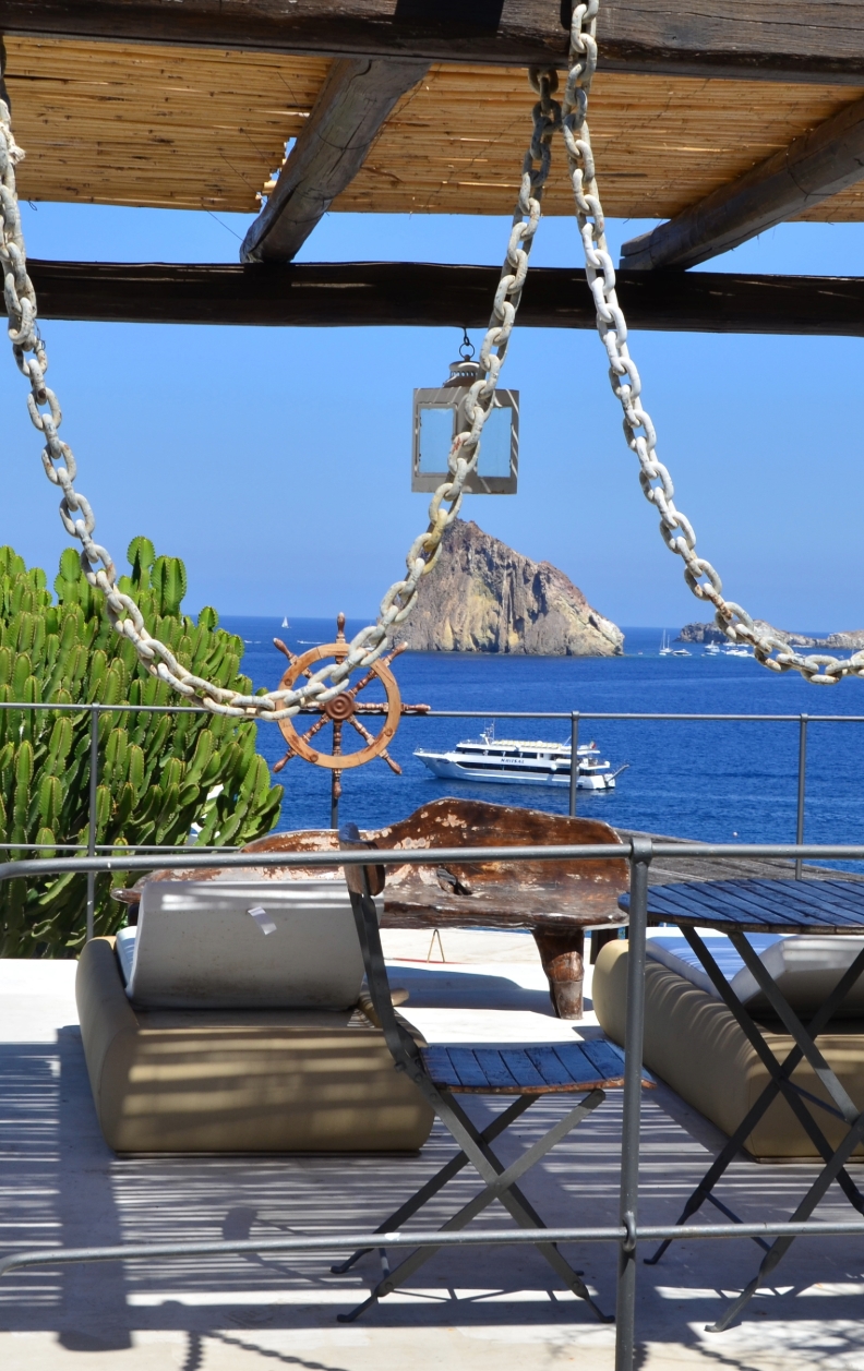 Sitzlounge mit Blick auf Strombolicchio bei Stromboli, aufgenommen von einer Terrasse mit Schiffsrad und Meerespanorama – Yachtcharter mit BlackSails-Yachting