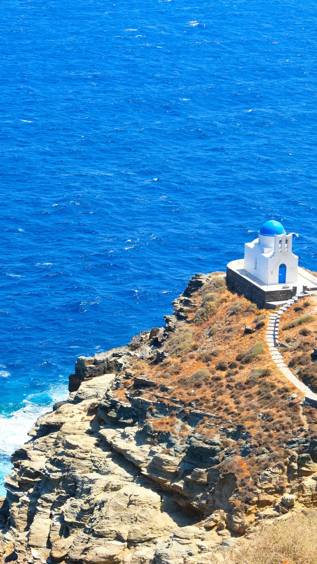 Weiße Kapelle auf einer Klippe mit tiefblauem, welligem Meer – typische Kykladenlandschaft, ideal für sportlichen Yachtcharter mit BlackSails-Yachting