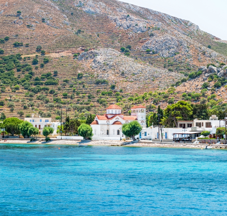 Weiße Kirche mit rotem Dach am Wasser auf einer Dodekanes-Insel, eingebettet in eine terrassierte Berglandschaft – typisch für Yachtcharter mit BlackSails-Yachting.