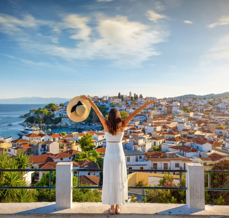 Frau in weißem Kleid blickt über eine Küstenstadt mit Yachthafen und hebt die Arme – Symbol für Freiheit beim Yachtcharter mit BlackSails-Yachting.