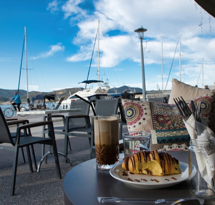 Tisch mit Kaffee und Gebäck in einem Hafenlokal, im Hintergrund Yachten, Berge und der Kai – perfekter Start beim Yachtcharter mit BlackSails-Yachting.