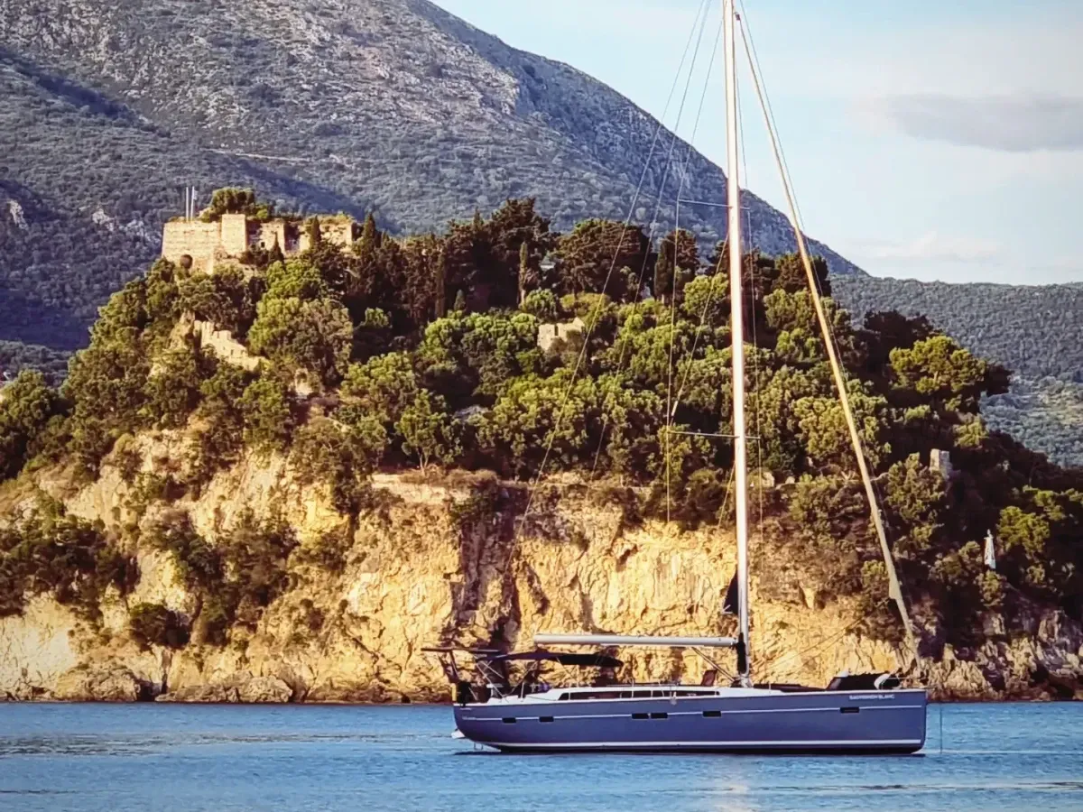 Eine stilvolle Segelyacht ankert vor den grünen Klippen von Parga, überragt von den Ruinen einer alten Festung. Im Hintergrund erheben sich bewaldete Berge, während der Himmel sich in sanften Blau- und Pastelltönen zeigt.