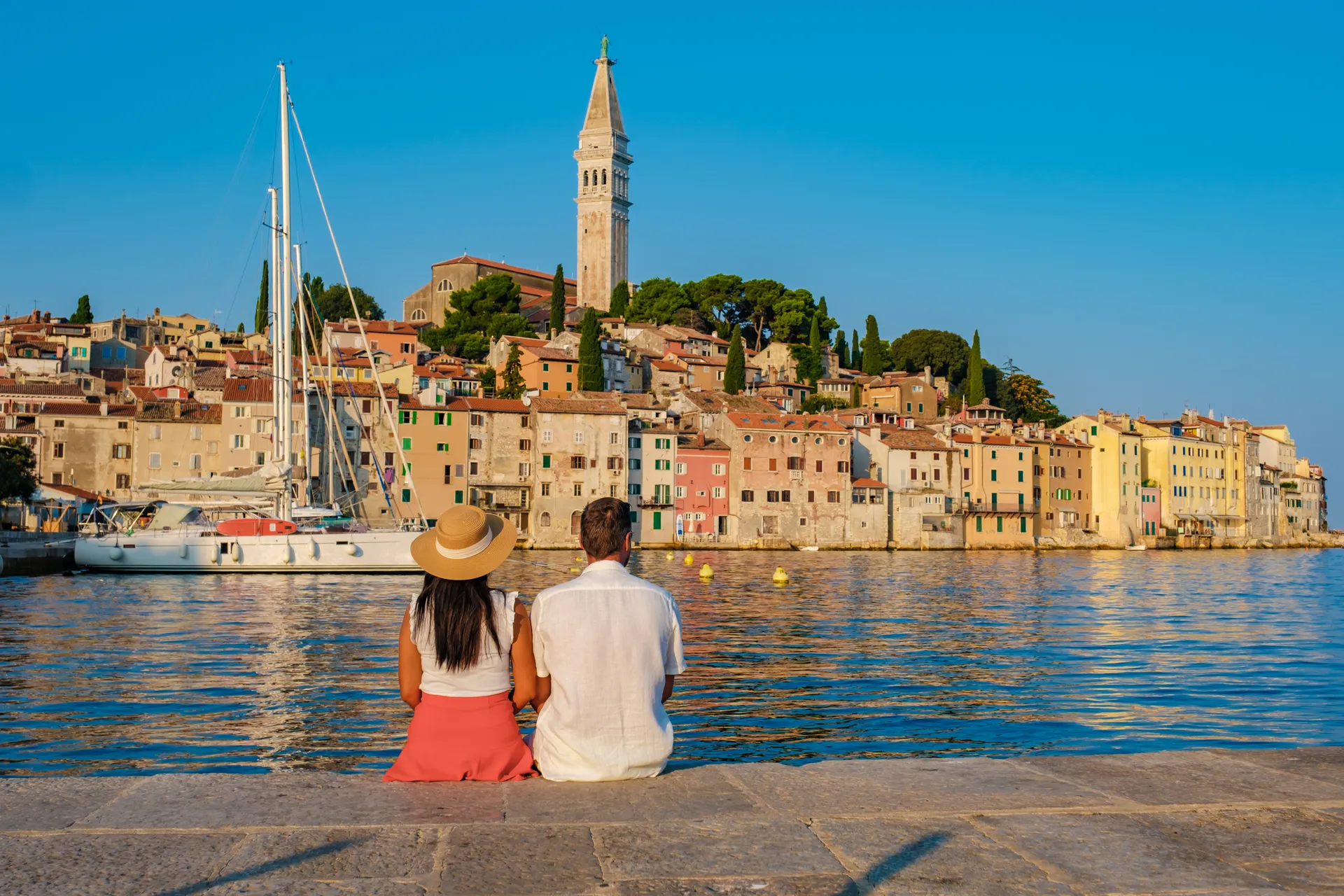 Ein Paar sitzt am Ufer in Rovinj, Kroatien, und genießt den Sonnenuntergang mit Blick auf die farbenfrohen Gebäude und eine Segelyacht im Hintergrund.