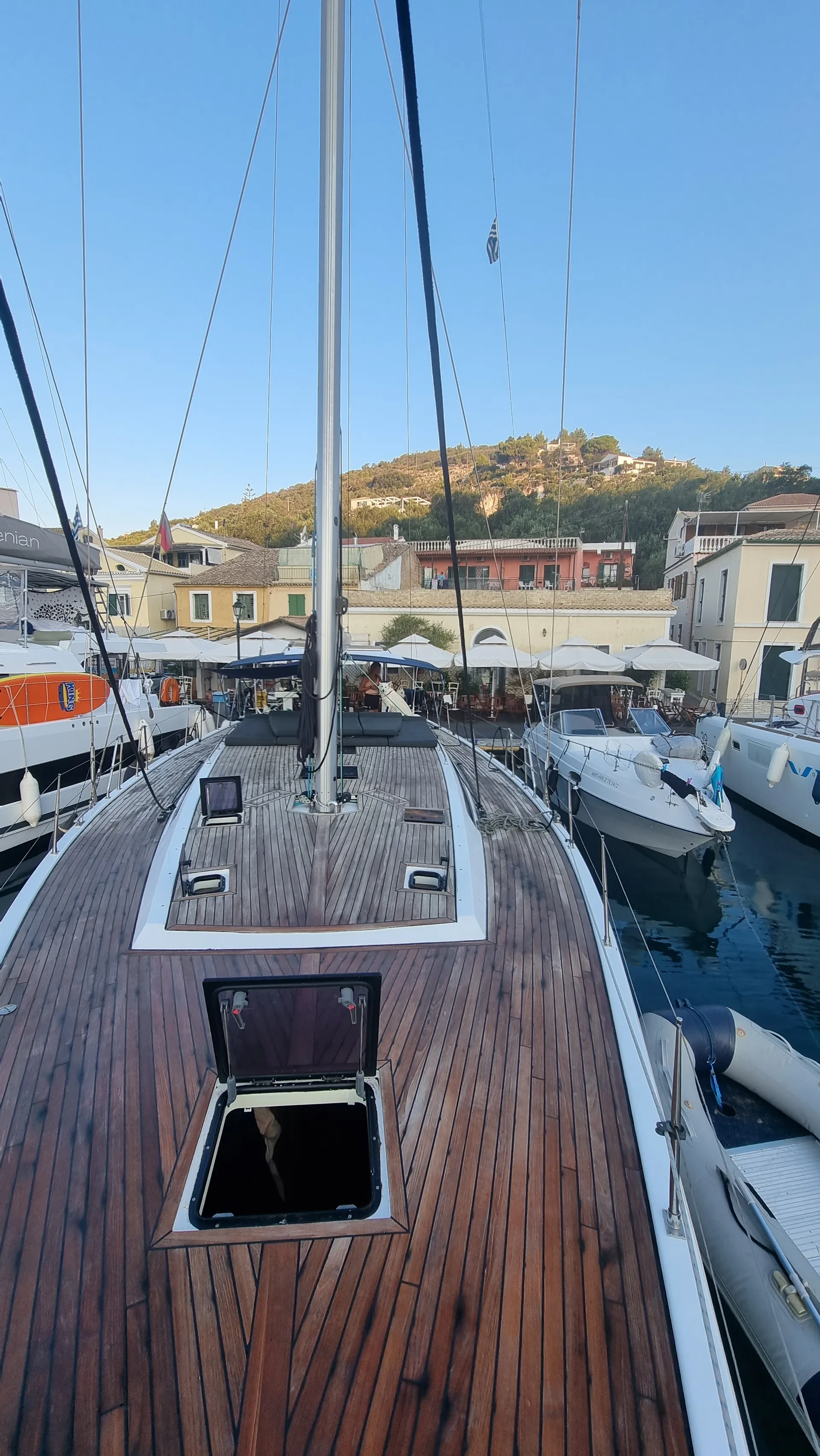 Blick vom Vordeck einer stilvollen Segelyacht im Hafen von Gaios, Paxos. Die Holzplanken glänzen in der Morgensonne, während sich bunte Häuser und Tavernen mit weißen Sonnenschirmen am Kai aneinanderreihen. Im Hintergrund ragen grüne Hügel über das malerische Dorf.