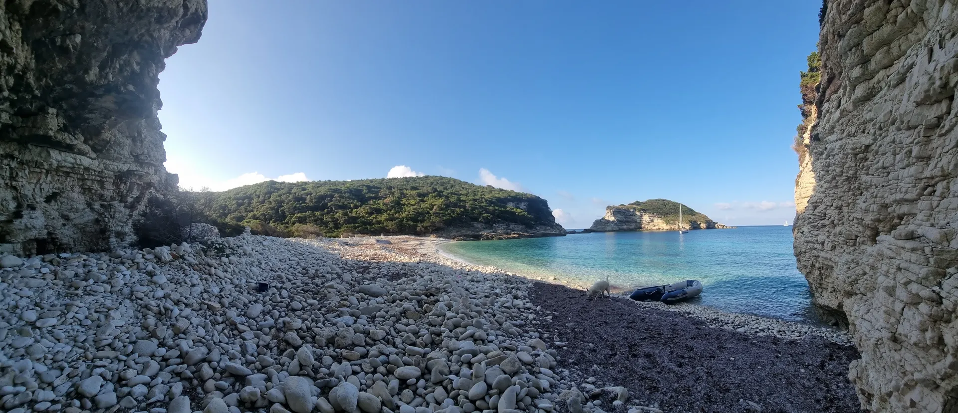 Ein abgeschiedener, steiniger Strand auf Antipaxos, umrahmt von hohen Felswänden. Ein Dinghi liegt am Ufer, während eine Segelyacht friedlich in der türkisblauen Bucht ankert. Ein Schaf grast am Ufer, während die Sonne den Himmel strahlend blau färbt.