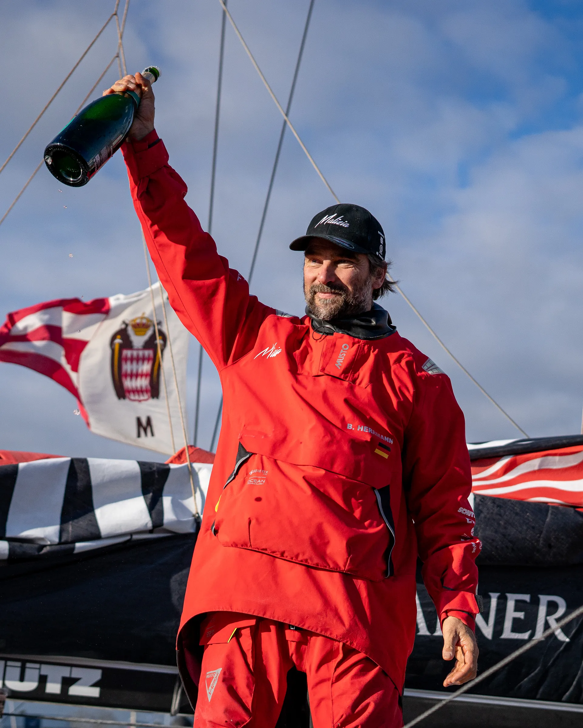 Boris Herrmann steht in roter Segeljacke auf der Malizia - Seaexplorer und hebt eine Champagnerflasche in die Luft – ein Moment des Triumphs nach einem epischen Rennen.