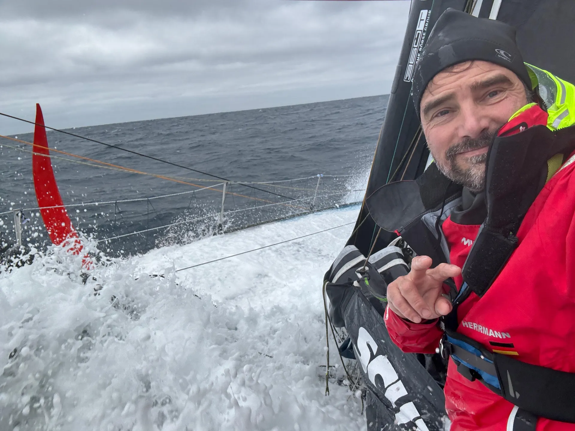Boris Herrmann an Bord der Malizia - Seaexplorer, in voller Segelmontur, während eine Welle über das Deck schlägt – pure Action auf der Vendée Globe.