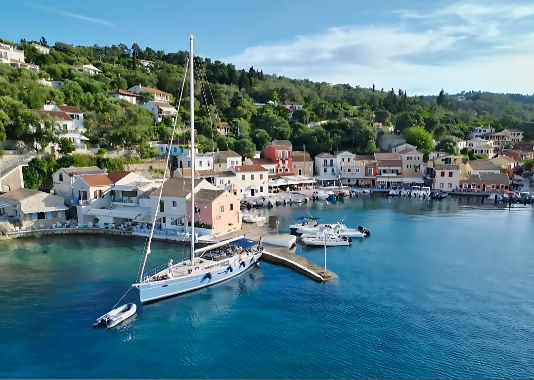 Eine stilvolle Segelyacht liegt längsseits am Pier im malerischen Hafen von Loggos auf Paxos. Im Hintergrund reihen sich charmante, bunte Häuser an die grüne Hügellandschaft, während das kristallklare Wasser in der Sonne glitzert.