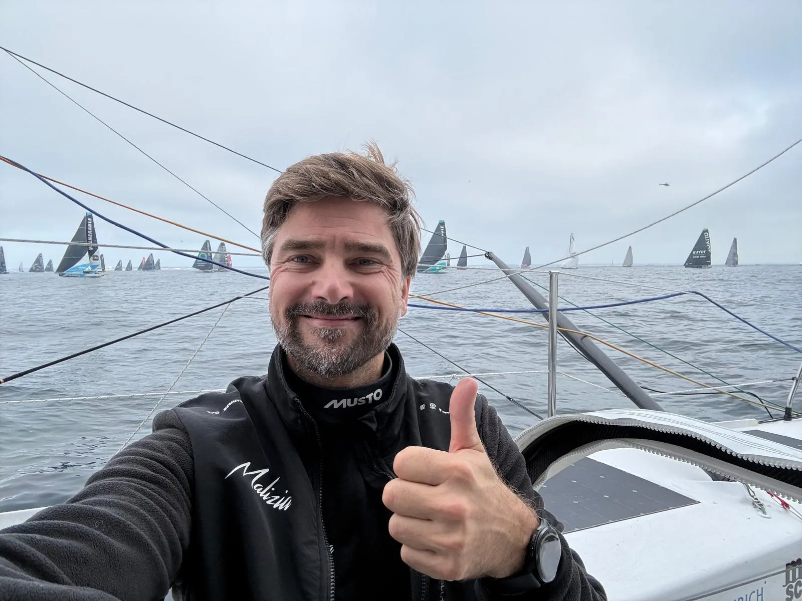 Boris Herrmann auf der Malizia - Seaexplorer zeigt einen Daumen hoch, während im Hintergrund mehrere IMOCA-Rennyachten auf dem offenen Meer zu sehen sind.