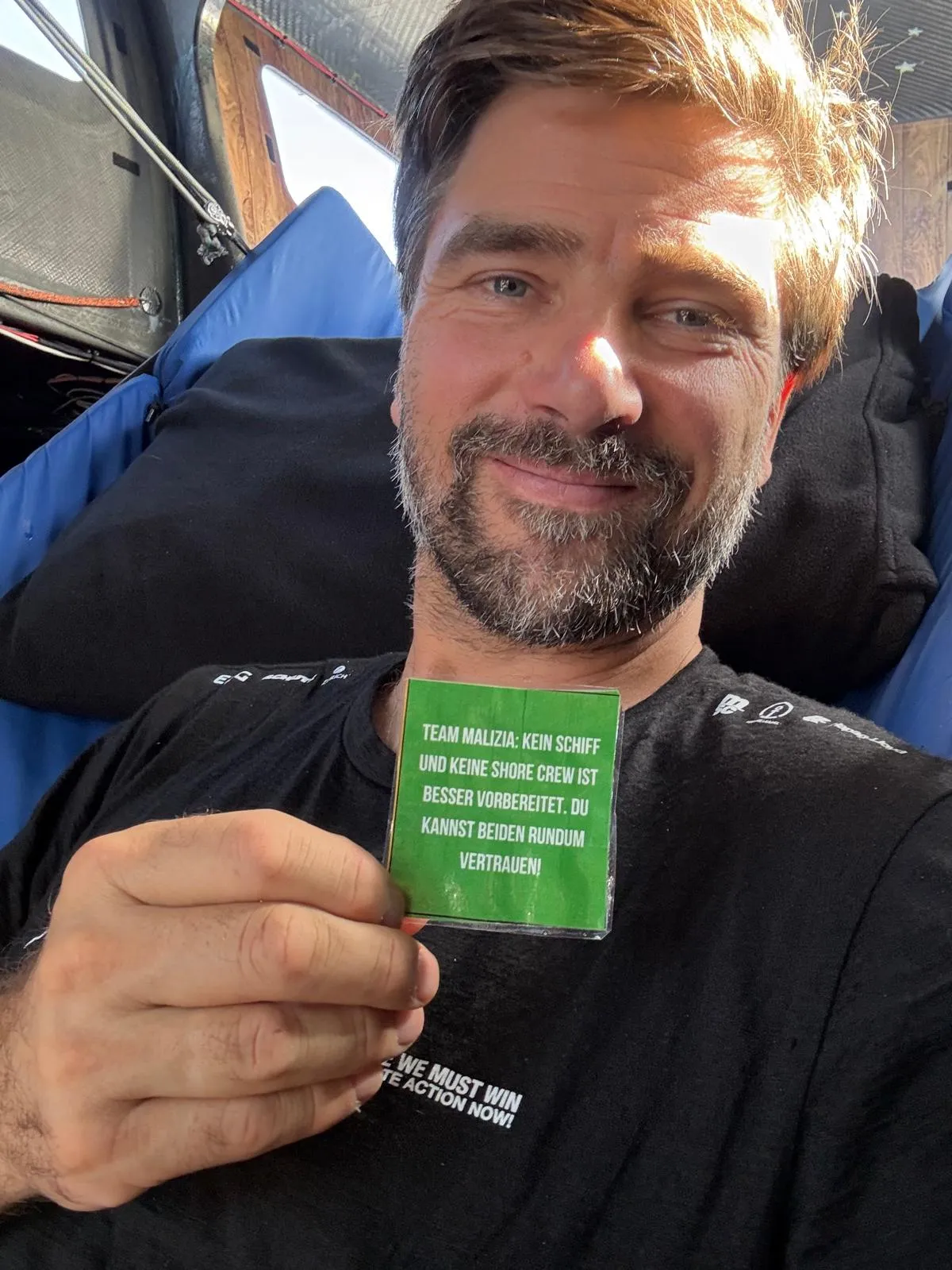 Boris Herrmann lächelt an Bord der Malizia - Seaexplorer und hält eine grüne Karte mit motivierender Botschaft über das Vertrauen in sein Team und sein Schiff.