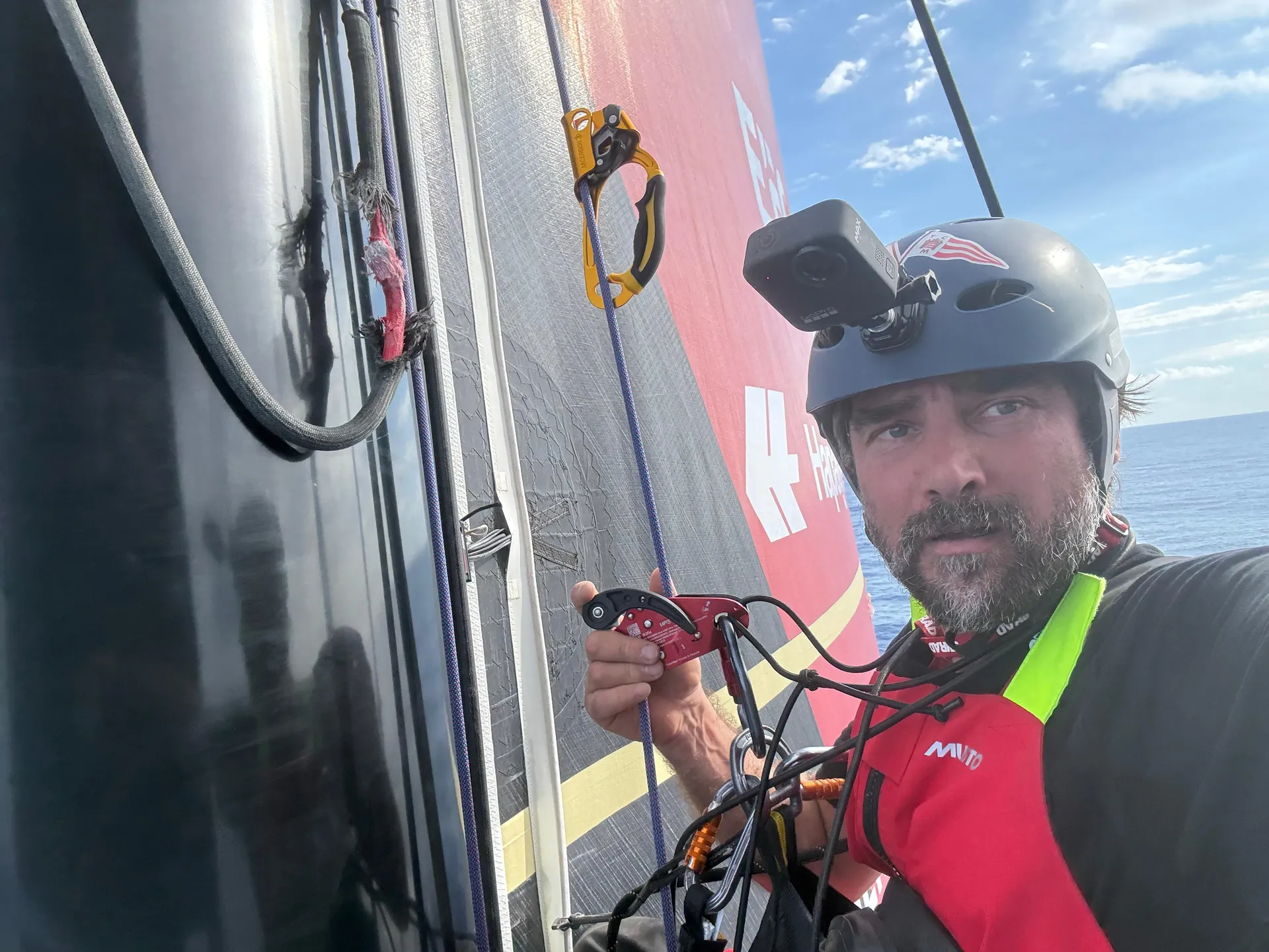 Boris Herrmann in Kletterausrüstung auf dem Mast der Malizia - Seaexplorer, während er eine beschädigte Leine inspiziert – ein riskanter Reparatureinsatz mitten auf hoher See.