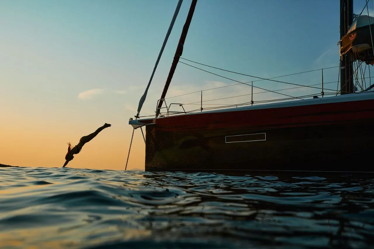Eine Frau taucht kopfüber ins Wasser neben der Kabuki Yacht, während die Sonne hinter dem Horizont versinkt – pure Freiheit auf einer Dufour 56 Exclusive.