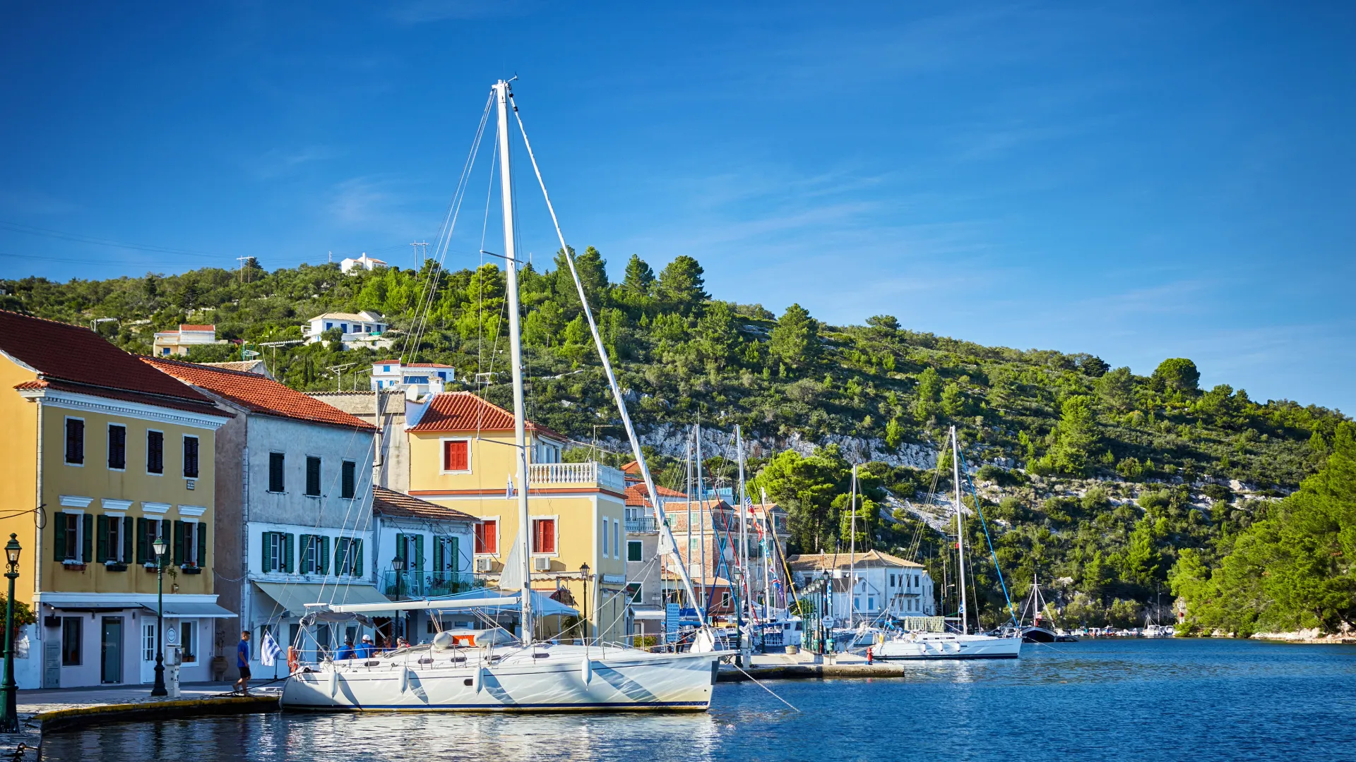 Segelyachten entlang der bunten Hafenpromenade eines griechischen Dorfes auf Paxos, umgeben von grünen Hügeln und klarem Wasser bei blauem Himmel.