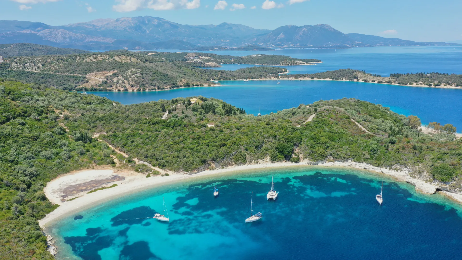 Drohnenaufnahme einer abgelegenen Bucht in Griechenland mit türkisblauem Wasser, weißen Segelyachten und dichter, grüner Vegetation im Hintergrund.