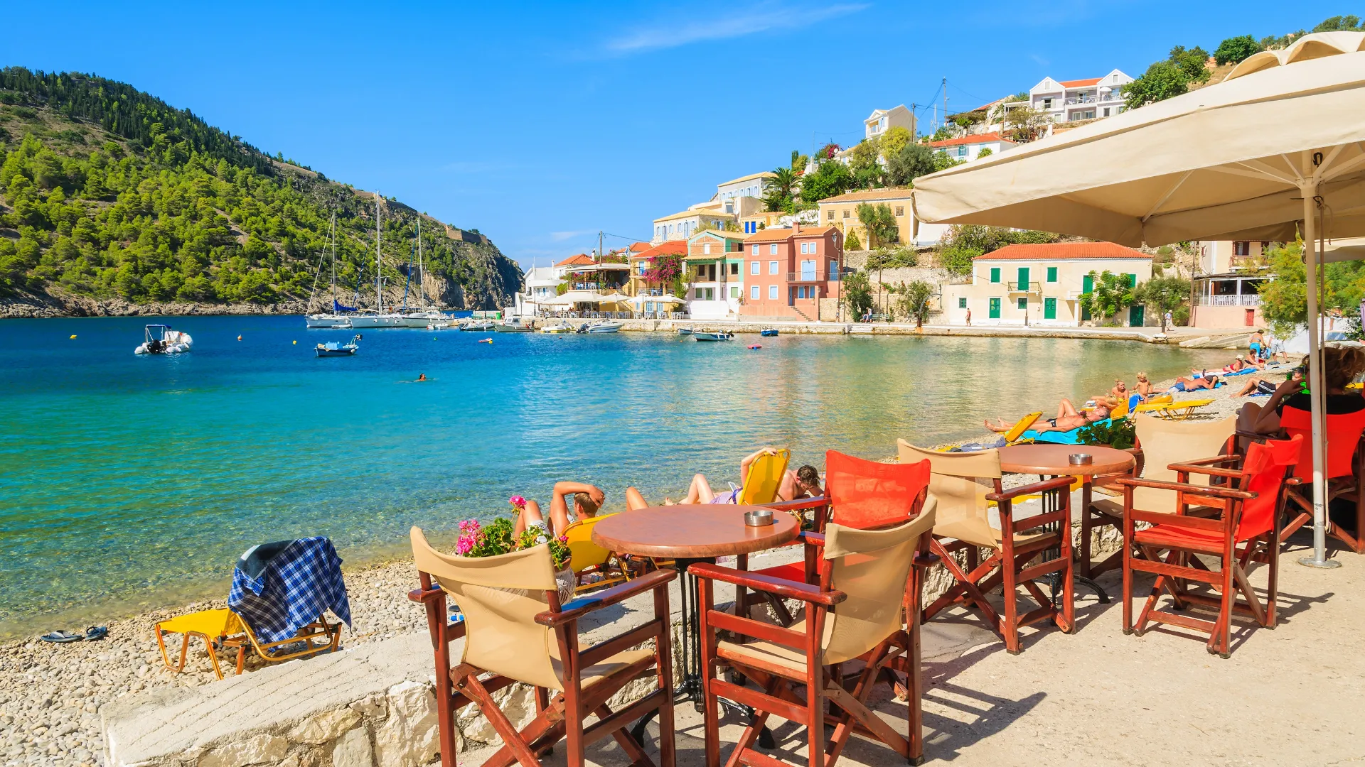 Café-Terrasse mit Blick auf das türkisfarbene Wasser einer Bucht auf Kefalonia, bunte Häuser, Liegestühle am Strand und Segelboote im Hintergrund.