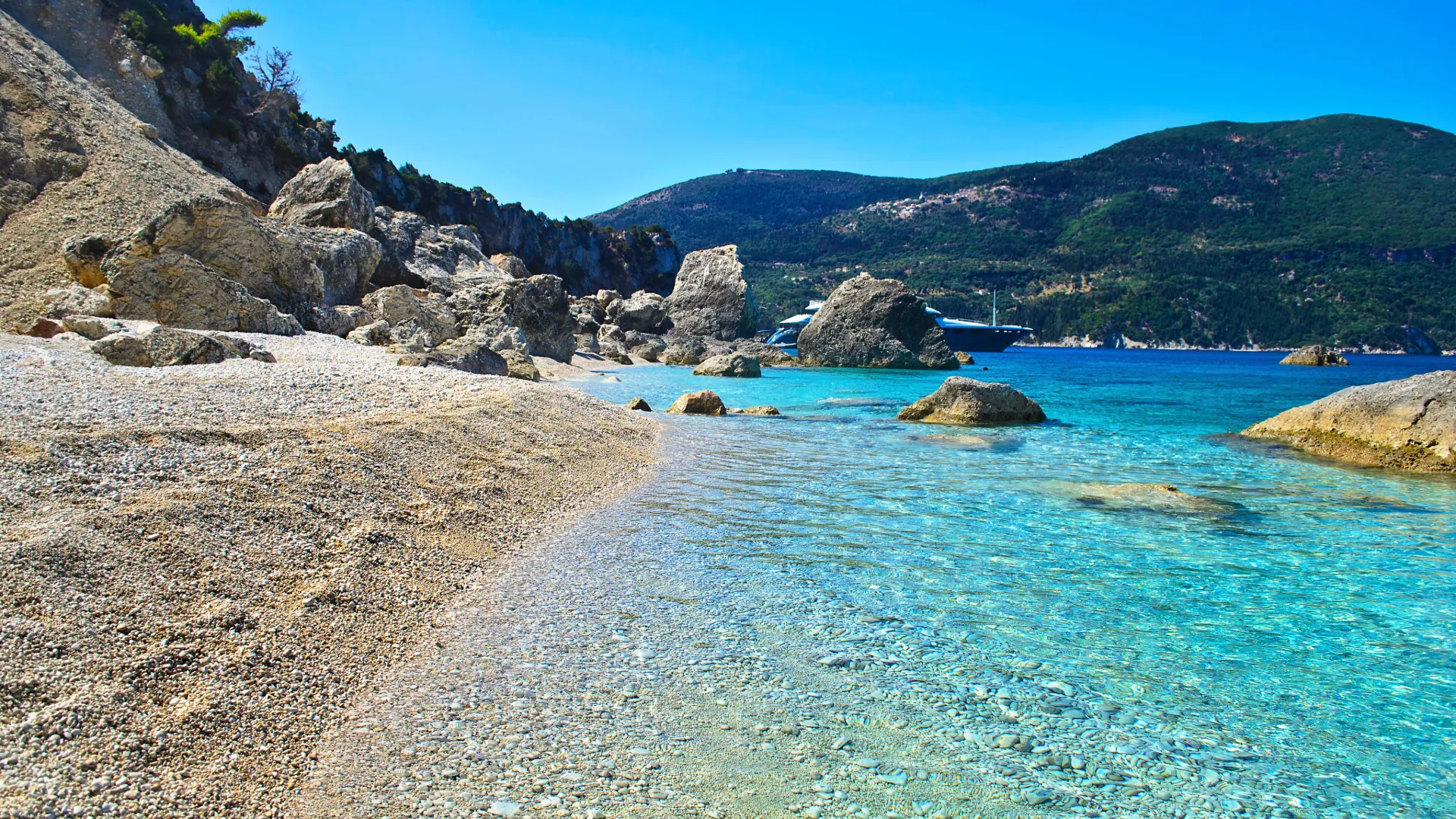 Kristallklares Wasser an einer felsigen Badebucht auf Kefalonia, Griechenland, mit großen Felsen, Kieselstrand und Yachten im Hintergrund vor bewaldeten Bergen.
