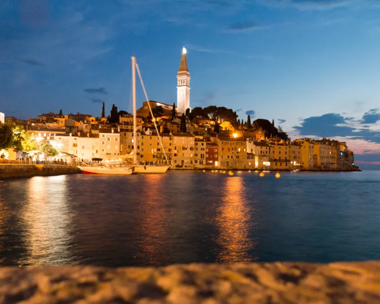 Segelyacht vor der Altstadt von Rovinj bei Sonnenuntergang, mit beleuchtetem Glockenturm der Kirche der Hl. Euphemia – BlackSails-Yachting Etappenziel in Kroatien
