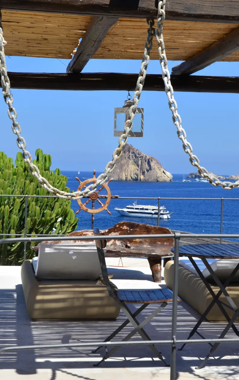 Sitzlounge mit Blick auf Strombolicchio bei Stromboli, aufgenommen von einer Terrasse mit Schiffsrad und Meerespanorama – Yachtcharter mit BlackSails-Yachting