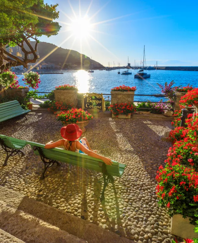Frau mit rotem Hut entspannt auf einer Bank in Marciana Marina mit Blick auf Segelboote und Sonnenuntergang – Yachtcharter mit BlackSails-Yachting auf Elba