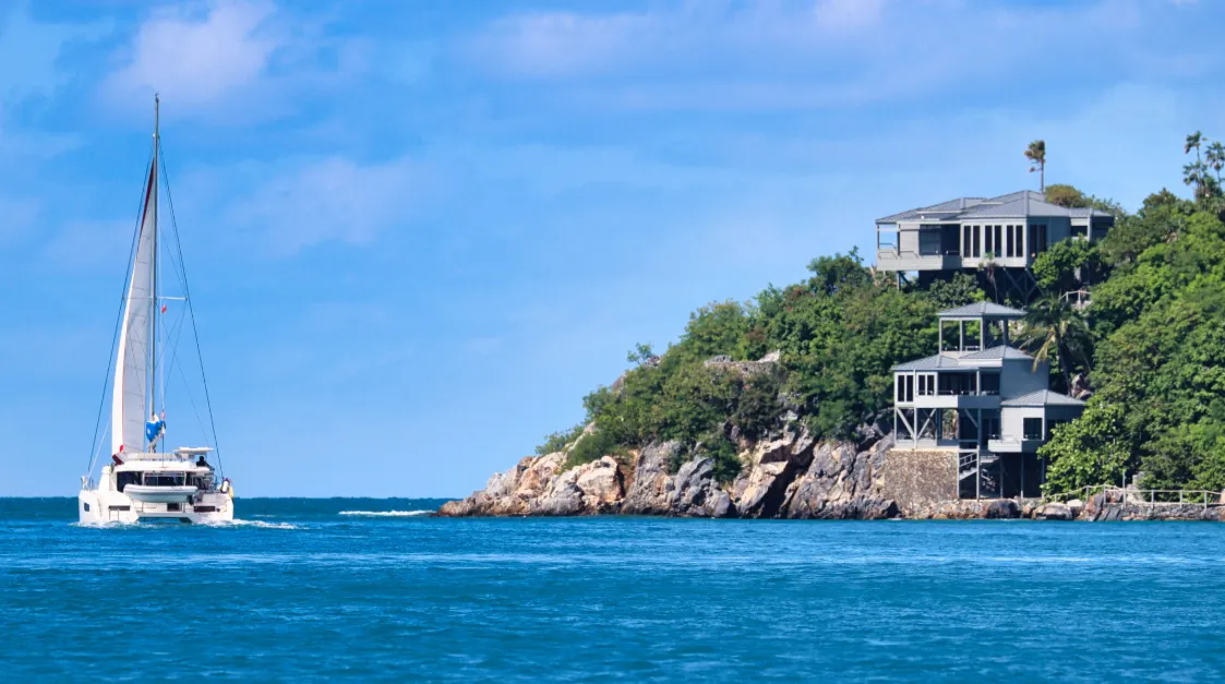 Segelkatamaran fährt entlang der Küste der British Virgin Islands mit modernen Villen auf felsigem, grün bewachsenen Hang im Hintergrund