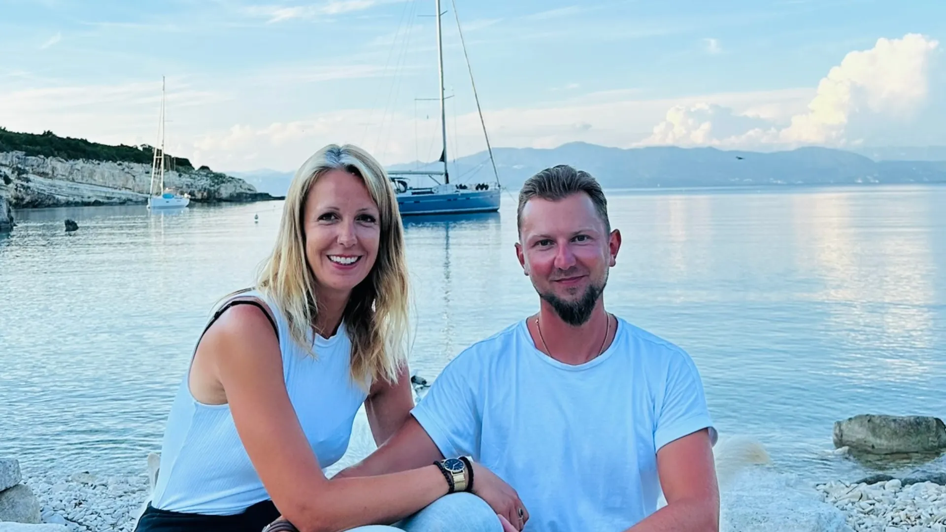 Laura und Daniel am Strand, im Hintergrund liegt ihr Segelboot vor Anker – ein Symbol für Partnerschaft, Freiheit und Gemeinschaft im Segeln.